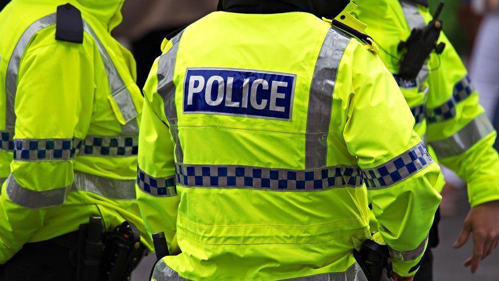 The back of of a police officer wearing a hi-vis jacket in the foreground with two other officers wearing hi-vis jackets in the background.