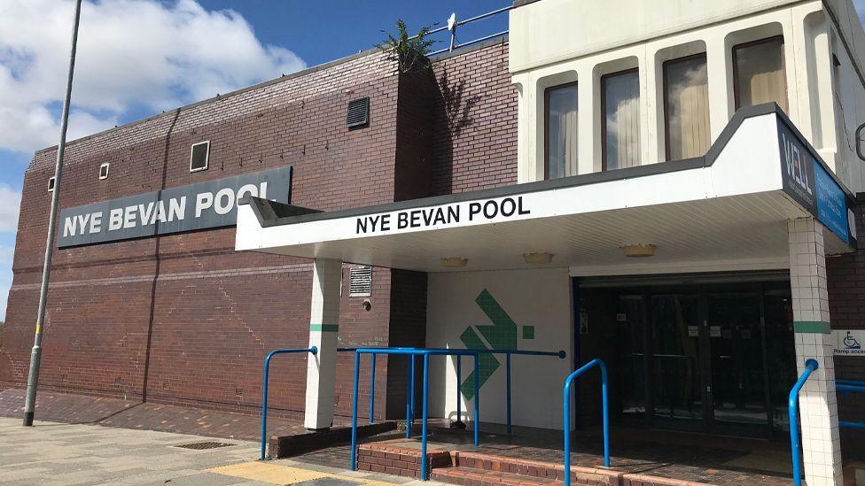 The outside of the Bevan Pool building. It has a large red brick square building to the left and a white overhanging porch to the right with blue access railings