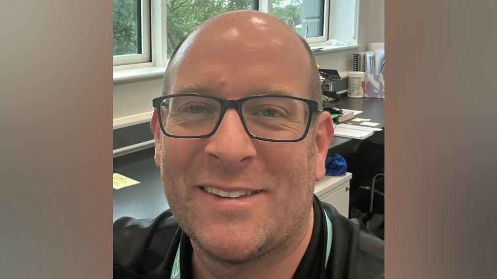 A man with a bald head and black glasses is smiling into the camera