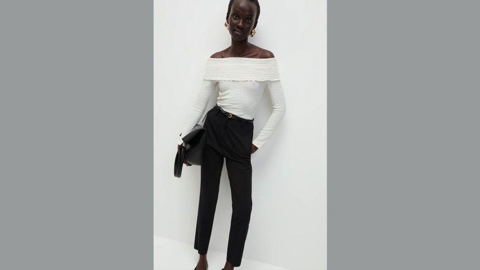 M&S model poses in white top and black trousers and leans against a white wall