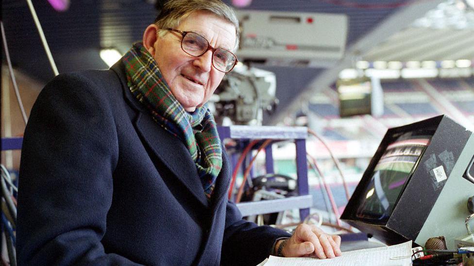 A man in dark blue coat and tartan scarf and glasses sits in front of a TV monitor with a commentary microphone beside it