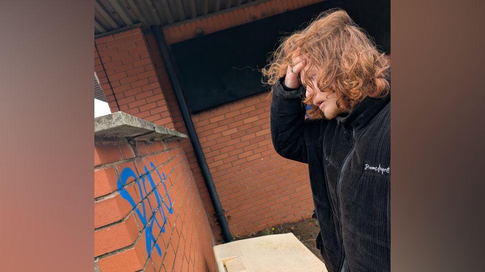 Theo Godfrey-Cass holds his hand to his head as he stares at the graffiti on the wall in disbelief. The blue tag has been sprayed onto the red brick.