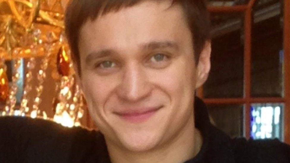 A man with close-cropped brown hair smiles in front of a large chandelier
