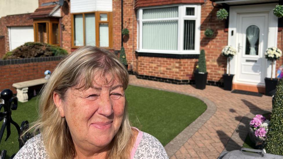 Elaine Mason outside her home in York. She is wearing a white top and stands in her garden which has artificial grass in front of her house