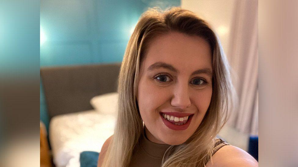 Lowri Puw  - a smiling woman with long blonde hair, wearing red lipstick. 