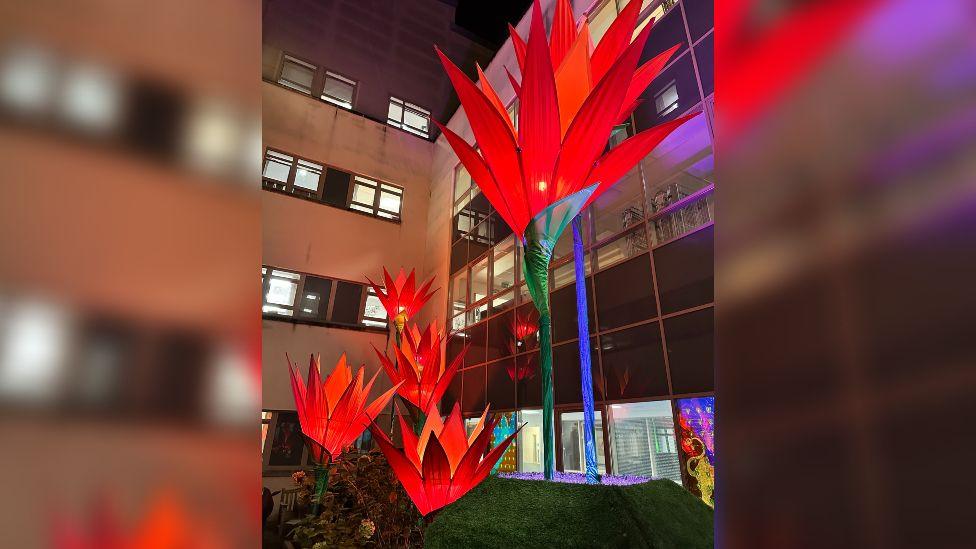 Five tall flower sculptures which are lit up in deep red hues at night outside the Royal United Hospital in Bath, where lights are on in the windows. 