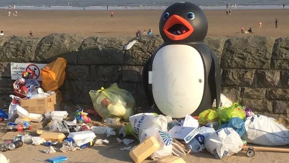 Rubbish left alongside Barry Island beach - plastic bags, boxes and fish and chip containers are strewn along the floor - next to a bin in the shape of a penguin.