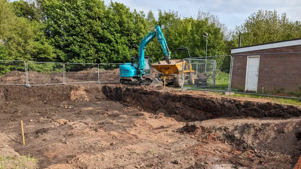 A large site in Carlisle which has been dug up by a blue digger and has been fenced off. There is a yellow pick-up truck where the digger has deposited the earth.