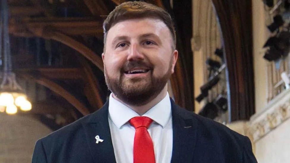 Head-and-shoulders image of Blackpool South MP Chris Webb smiling, wearing a dark suit, white shirt and red tie.