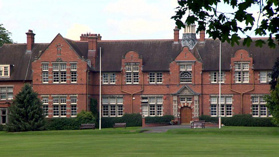 A large two-storey red brick building with a large lawn in front. Two flag poles can be seen in front of the building.