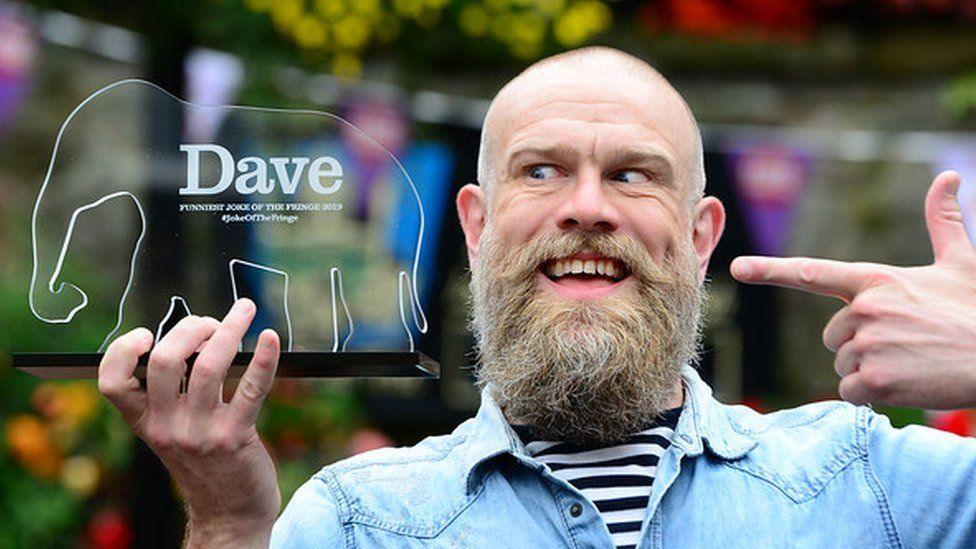 Olaf Falafel, a man with a big beard wearing a denim shirt with a striped t-shirt underneath. He is pointing towards a trophy shaped like an elephant and pulling an odd expression.