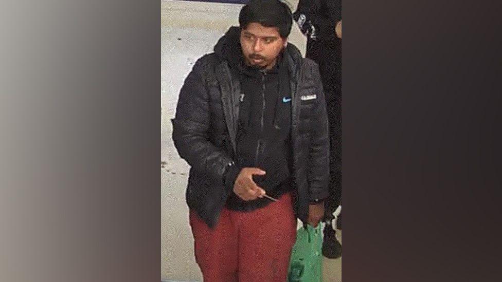 Missing man Yusef is seen on a CCTV camera walking and looking slightly to his right. He has thick dark smooth hair and a moustache and goatee beard. He is wearing a black hoodie and open black puffa style jacket, and red trousers. He is carrying a green carrier back and there is the elbow of another person to his left. 