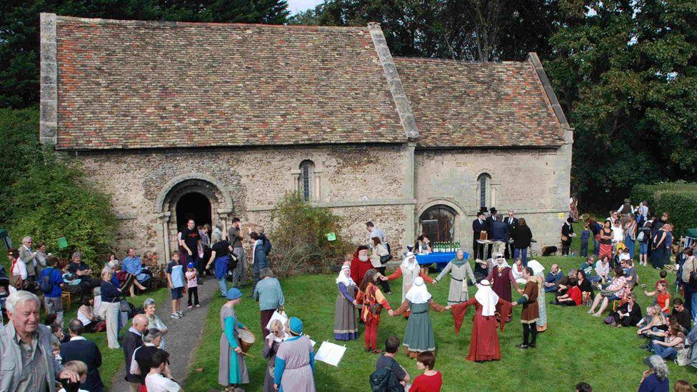 A small single storey medieval stone church with a tiled roof called the Leper Chapel, in Cambridge. Its arched door is on its left and there are three arched windows, two very narrow. In front of it is a path going up to the door and grass. There are many people milling around, most watching medieval re-enactors, a group of whom are holding hands and dancing