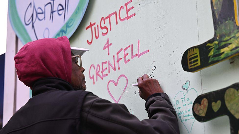 A person wearing a cap and glasses with their hood up draws on a wall that says 'justice 4 Grenfell' next to a picture of a heart, in London on 14 June