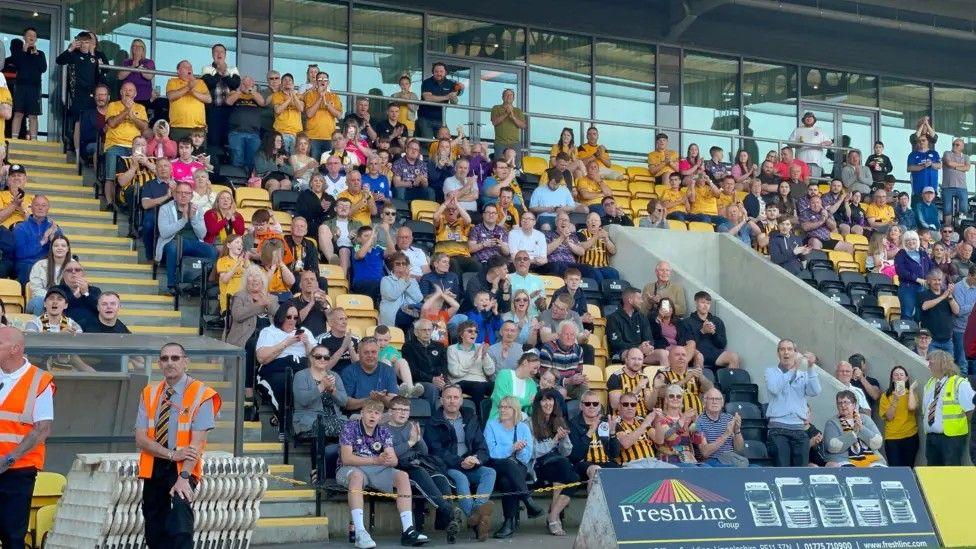 Fans, many wearing yellow and black football shirts, sitting in a stand at a football ground. There are some officials dressed in orange hi-vis vests in the foreground, along with some advertising hoardings.