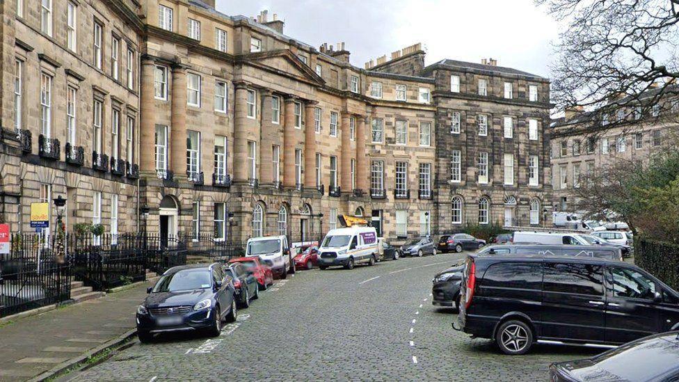 Moray Place sweeeps round to the right. It has beautifully grand sandstone buildings. There are cars parked on its cobbled road.