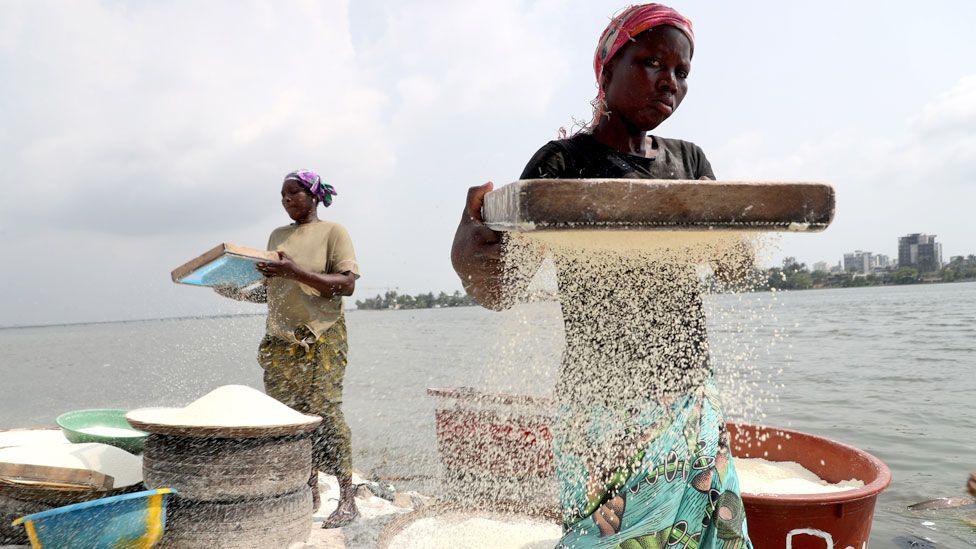 Women stand by the sea and sieve cassava grains into large pots. Abidjan's skyline can be seen in the background - Thursday 11 December 2024