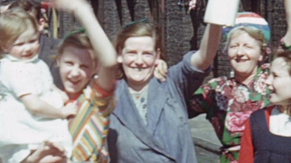 Grainy photo of three adult women, one girl on the righthand side and a baby in a white dress. The women are all smiling and waving. The woman on the left is wearing a multicoloured yellow and brown top. The woman in the middle in wearing blue overalls. The older woman in the back is wearing a hat and a green top with pink roses on it. 
