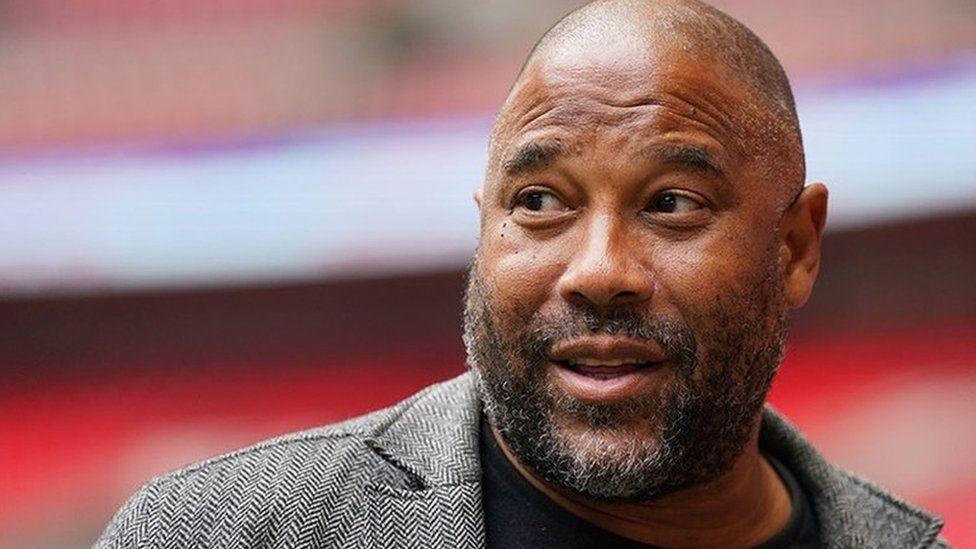 John Barnes, who has a bald head and black and grey stubble, looks at the camera wearing a grey coat