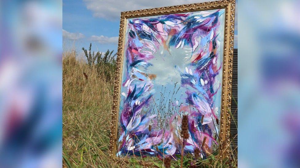 The mirror photographed propped up in a grassy field with blue sky behind it. It's in a gilded metal frame and is painted around the edges with an abstract flower pattern in shades of blue, purple, pink and white.