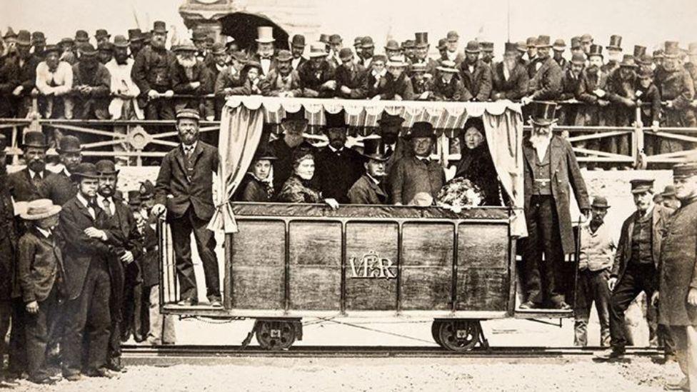 A black and white image of a large group of men. There is a handful of men inside a carriage on the Volk's Railway. 