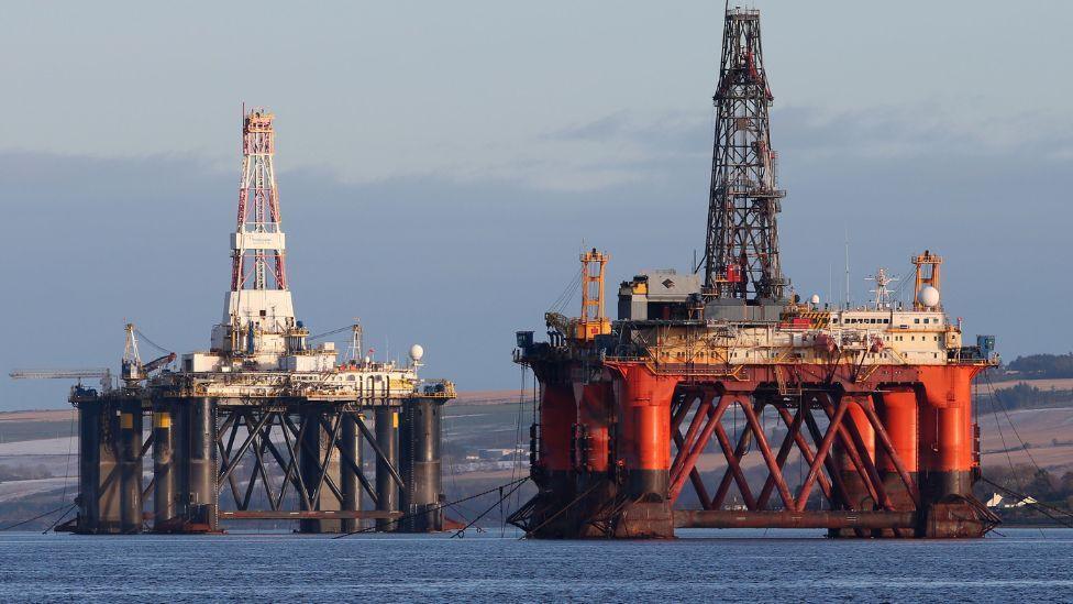Two oil metal oil platforms, one red and the other black, in a flat blue sea, with low hills in the background.