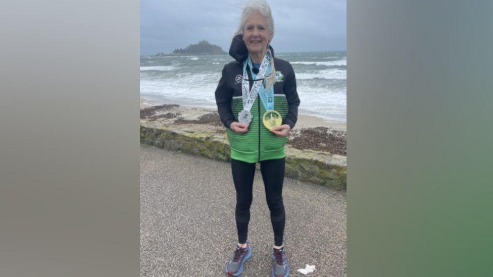 Andrea Simmons stands in front a beach on a choppy day as waves crash onto the sand. The 71-year-old has a Hayle Runners shell suit top on and two large medals around her neck.