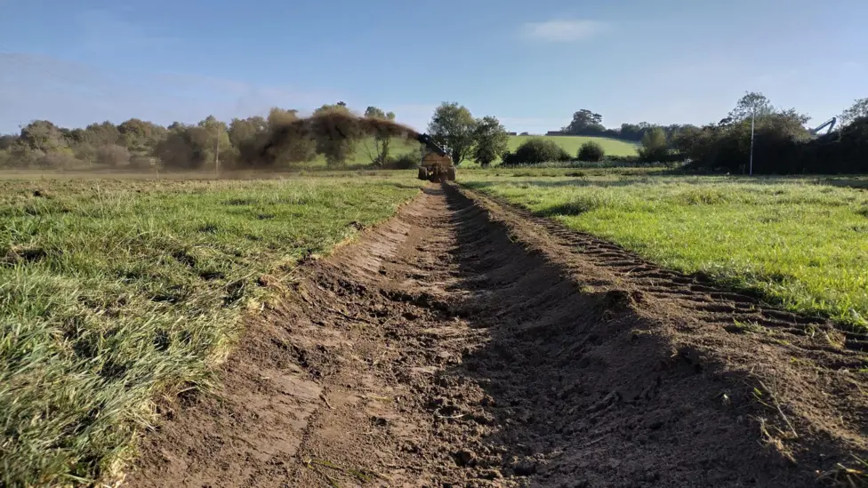 Image shows large brown channel carved in a green, grassy, field with a tractor in the distance.