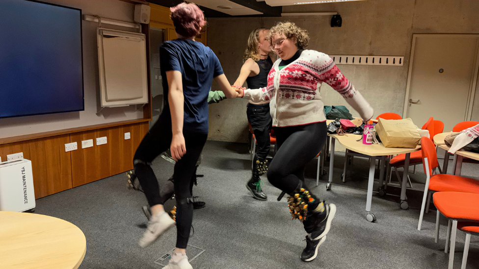 Four people, with bells on their ankles, dancing in a circle inside a university classroom