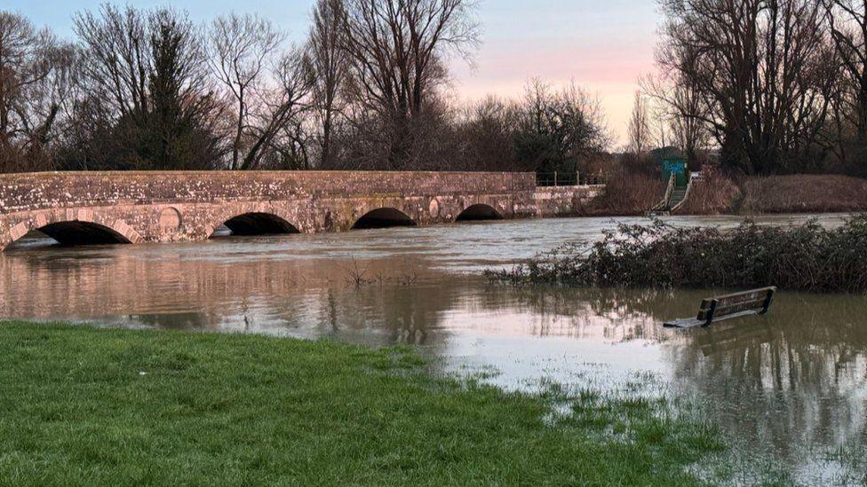 Grass areas with bench underwater and river water reaching high up on four arches of a brock bridge spanning the water.