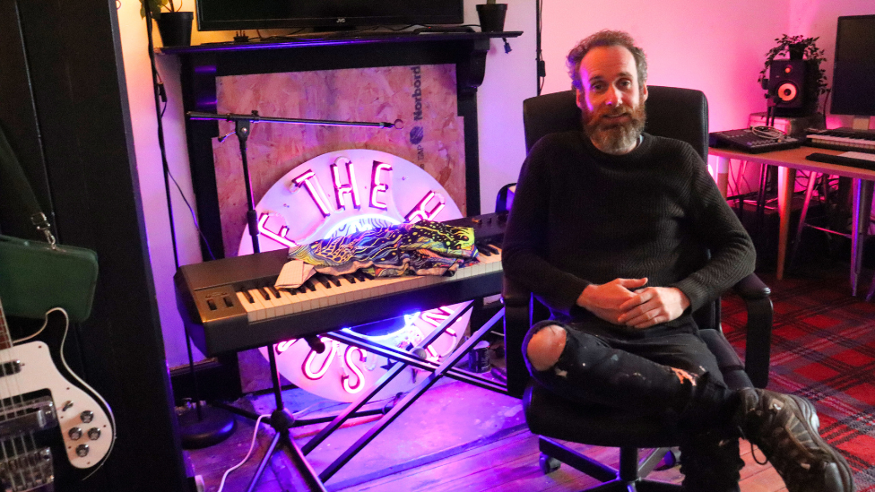 Mick McCullagh in a black jumper and ripped jeans, sat in front of a keyboard and a neon purple light, a guitar and a computer can also be seen in the background