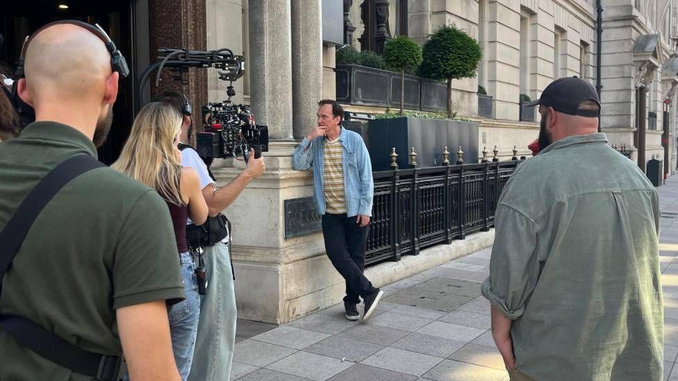 Quentin Tarantino is seen wearing a blue jacket and white t shirt, leaning against the wall of a hotel. In front of him is someone with a camera, while support crew are also standing around