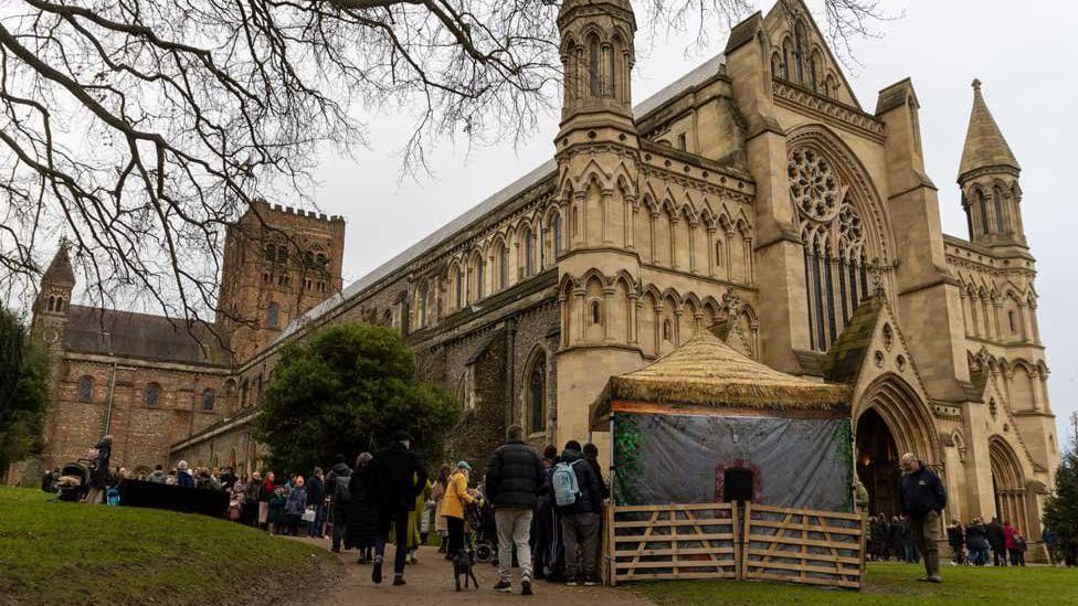 The west end of St Albans' Norman cathedral has a wooden barn outside the entrance, with many people visiting it.