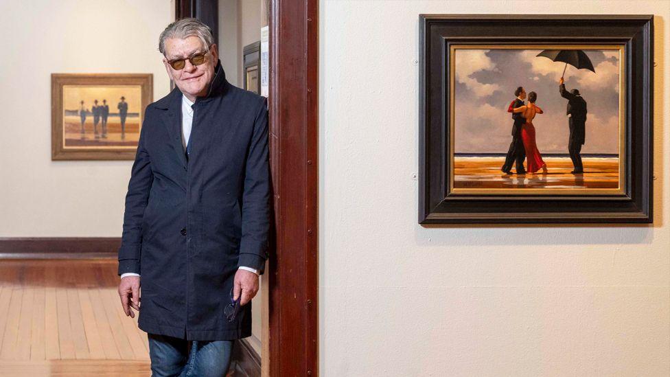 Jack Vettriano standing in a gallery, leaning against a wall and wearing a blue coat and jeans. His pictures are hanging on the walls behind and to the right of him.