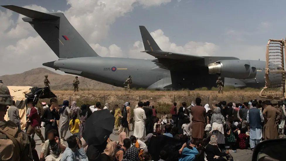 A crowd of adults and children wait on the tarmac at an airport. In the background, a large military transport plane can be seen, guarded by British soldiers. 