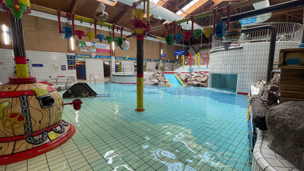 The swimming pool at Leisure World. The water is blue and runs over a white tiled floor. There are buckets that pour water over people suspended from a long yellow pole. There is a covered area with a waterfall in front of it. There are entrances to the changing rooms in the background and a blue slide.