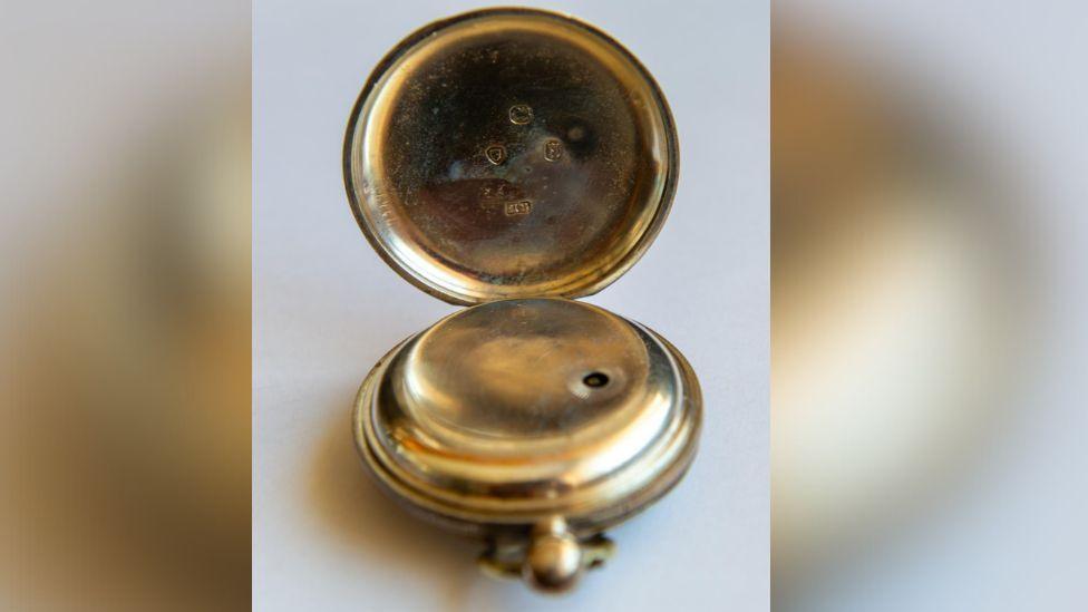 A close-up shot of the gold pocket watch. The watch is open and has several small engravings inside the case. 