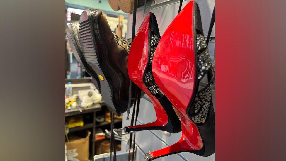 Red and black stilettos hanging on a wall next to a pair of walking boots.