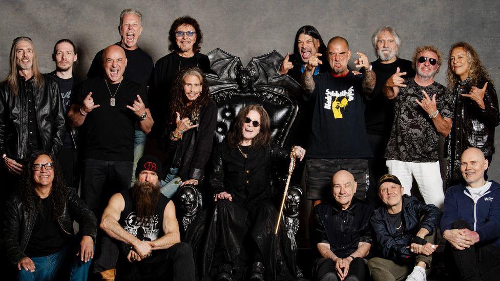 Ozzy Osbourne seated on a black throne in the centre, with 16 other musicians standing and sitting either side of him, in a photo taken before the gig