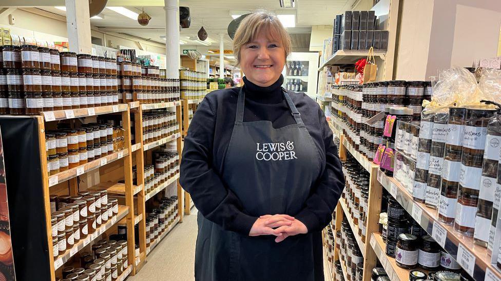 A woman in a black top and black "Lewis & Cooper" apron is standing in front of shelves with jars of products 