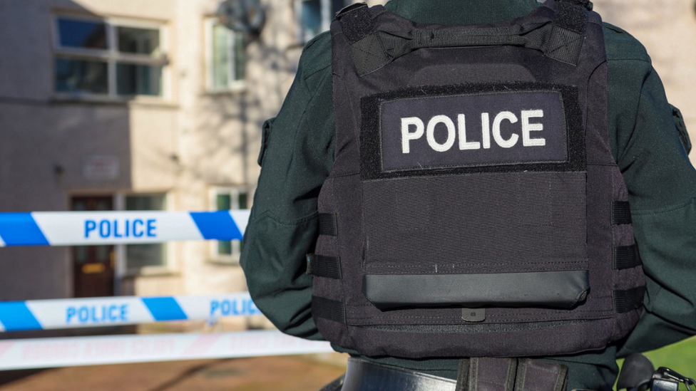 A a police officer with a dark green coat on and a black 'police' bullet proof vest and a black cap with 'police' on the back of it. In front of him are flats in a residential area and a blue and white 'police line do not cross' tape.