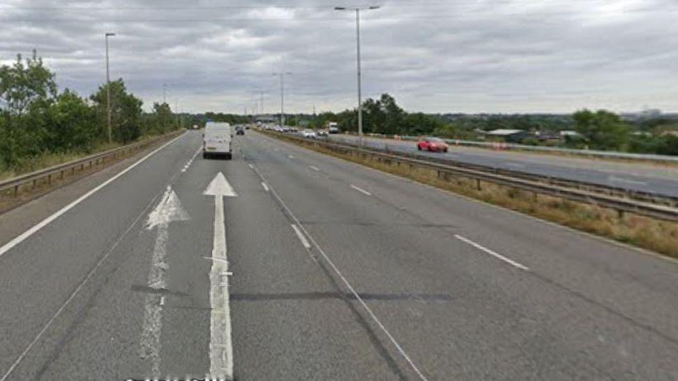 The M40 and the slip road on the left which leads to the A40. There are three lanes of motorway on each side plus the hard shoulder/slip road. A white van is seen from behind and on the road are two arrows – the left one marking the slip road exit junction. It is daylight