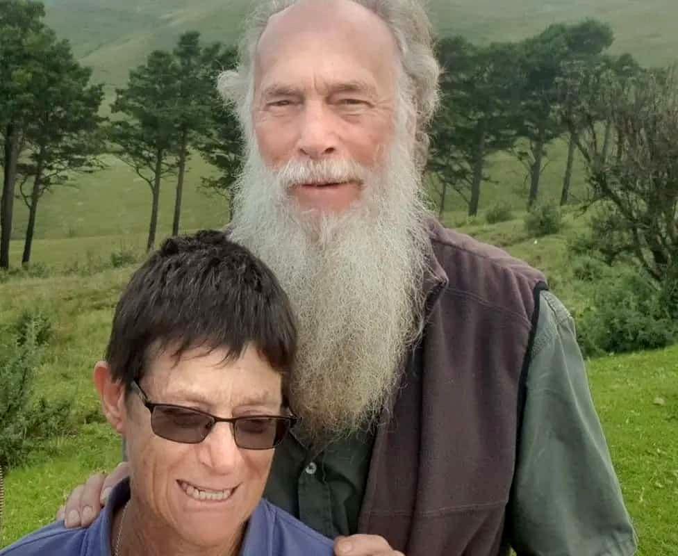 Rodney Saunders stands behind Rachel. Rodney has a long grey beard while Rachel has dark hair and is wearing sunglasses