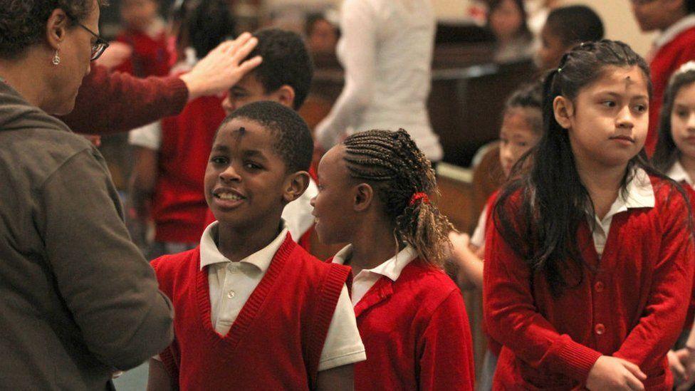 Children wearing school uniform are shown with black ash in the shape of a cross on their forehead