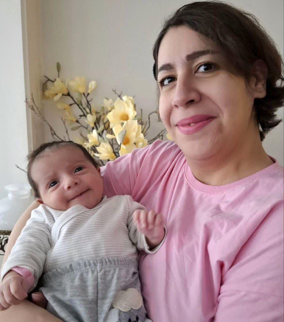 Photograph of Rezvaneh holding her baby daughter. Rezvaneh has short to medium-length dark hair, is smiling and is wearing a light pink top. Her daughter has dark hair and is wearing white and grey striped top and grey trousers. There are yellow flowers in the background.