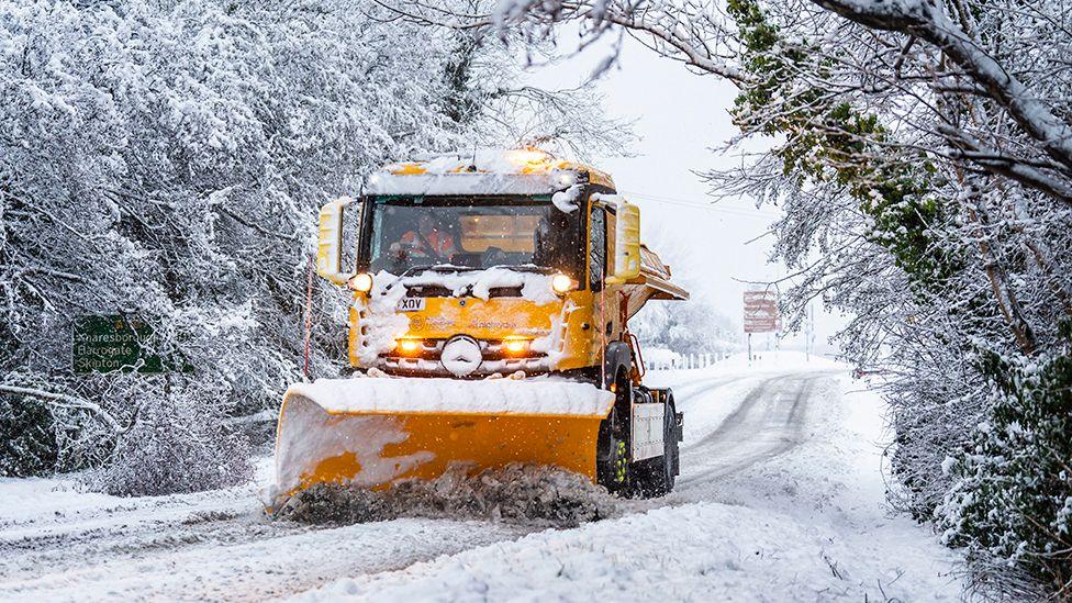 A gritter clearing snow on a road surrounded by snow-laden trees