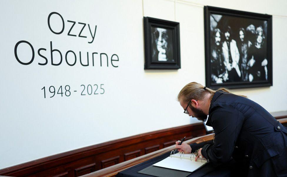 A man with a dark suit, ponytail and beard signing a book of condolence in front of a wall with large wording saying "Ozzy Osbourne 1948-2025"