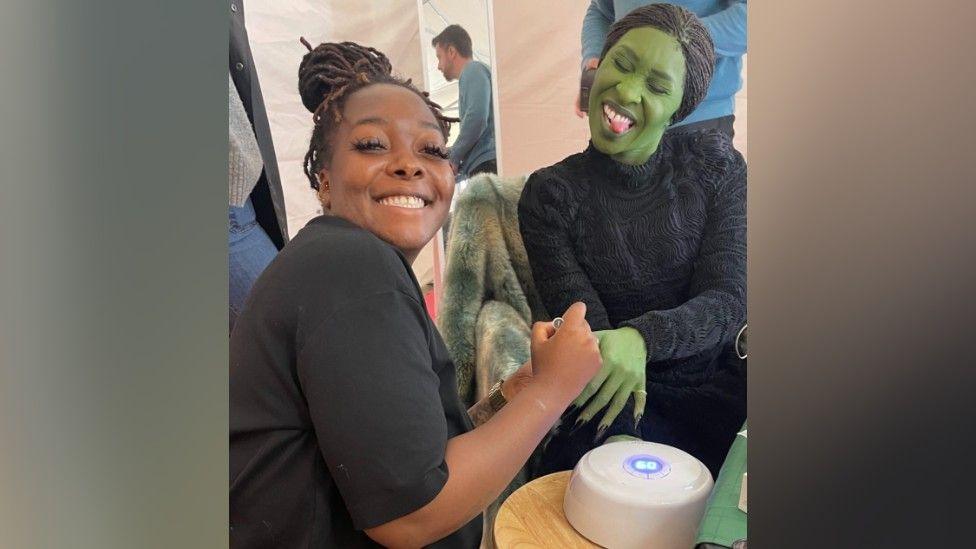 Shea Osei has her hair tied up and is smiling at the camera. She is wearing all black and is sat doing Cynthia Erivo's nails. Cynthia Erivo is also wearing black and has her body and face painted green. She has her eyes closed and is sticking her tongue out to the camera. The pair are sat around a wooden table with a gel lamp on it.