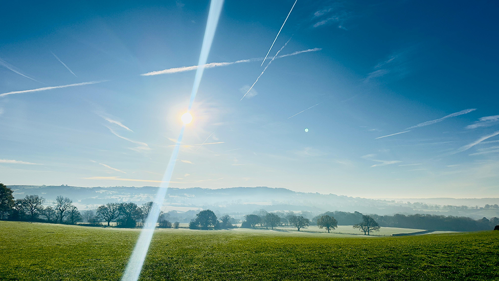 The sun shines brightly over a field and hillside in Leek. There is a mist in the distance, partly shrouding the tree-line and hills beyond