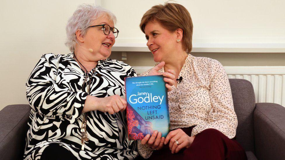 Janey Godley and Nicola Sturgeon sitting on a sofa holding up Janey's autobiography